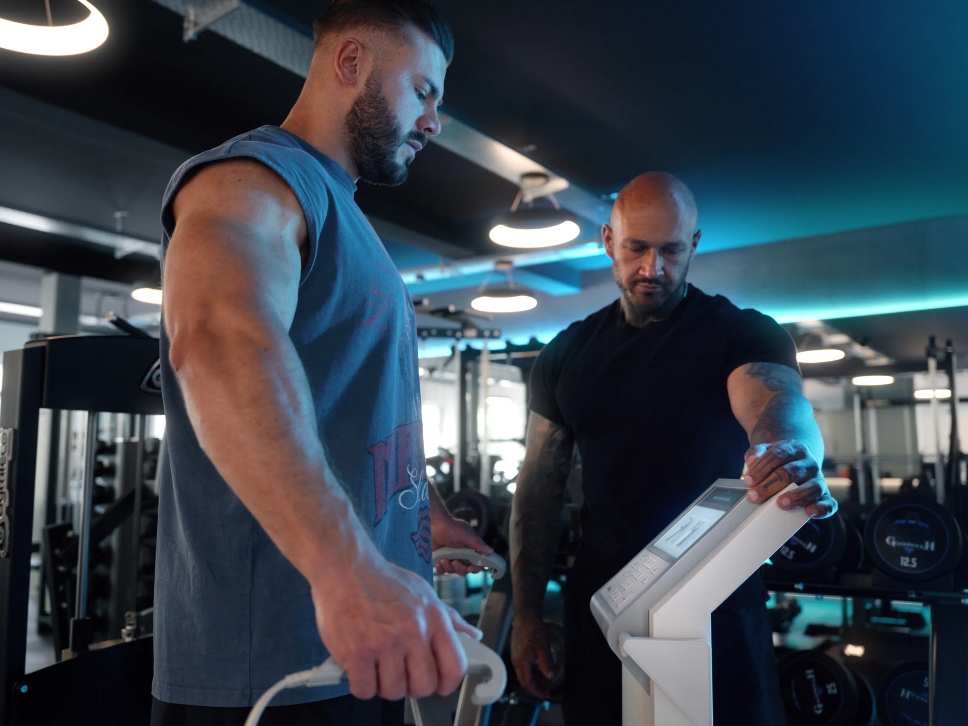 Zwei muskulöse Männer im Fitnessstudio bedienen ein elektronisches Trainingsgerät.