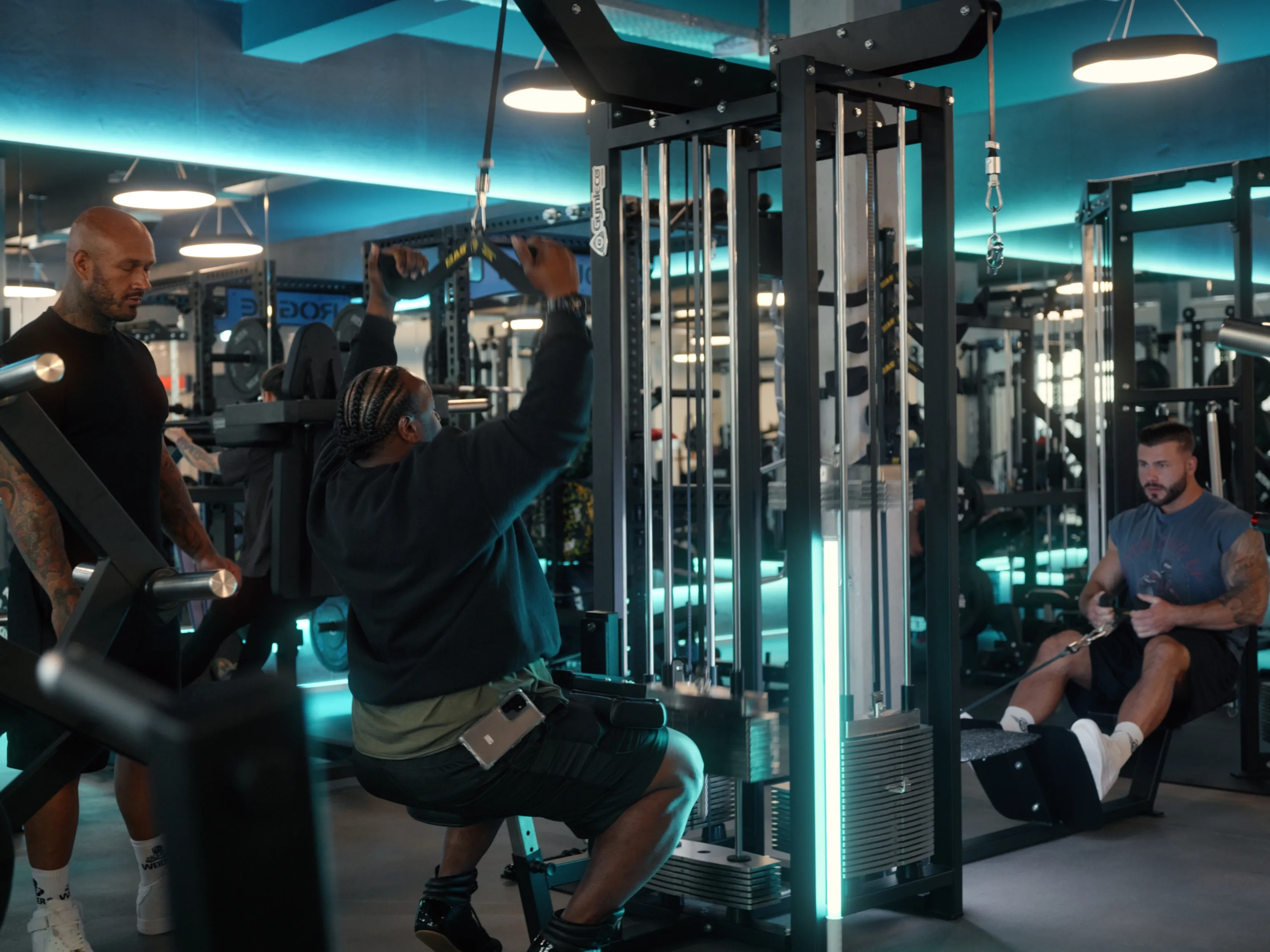 Zwei Männer trainieren an Kabelzugmaschinen im Fitnessstudio mit einem Trainer, der zuschaut.