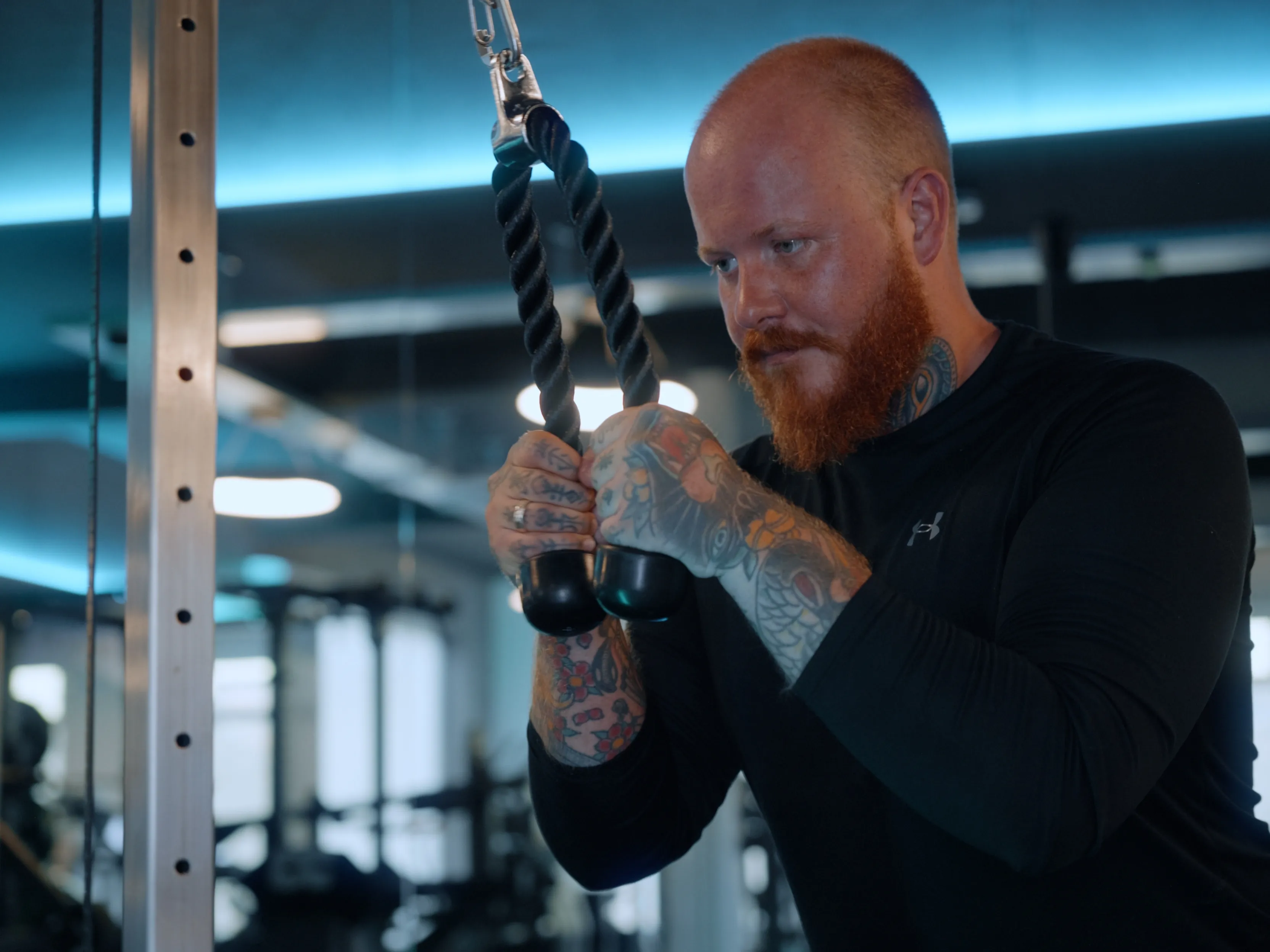 Tattooierter Mann mit rotem Bart trainiert mit einem Seilzuggerät im Fitnessstudio.