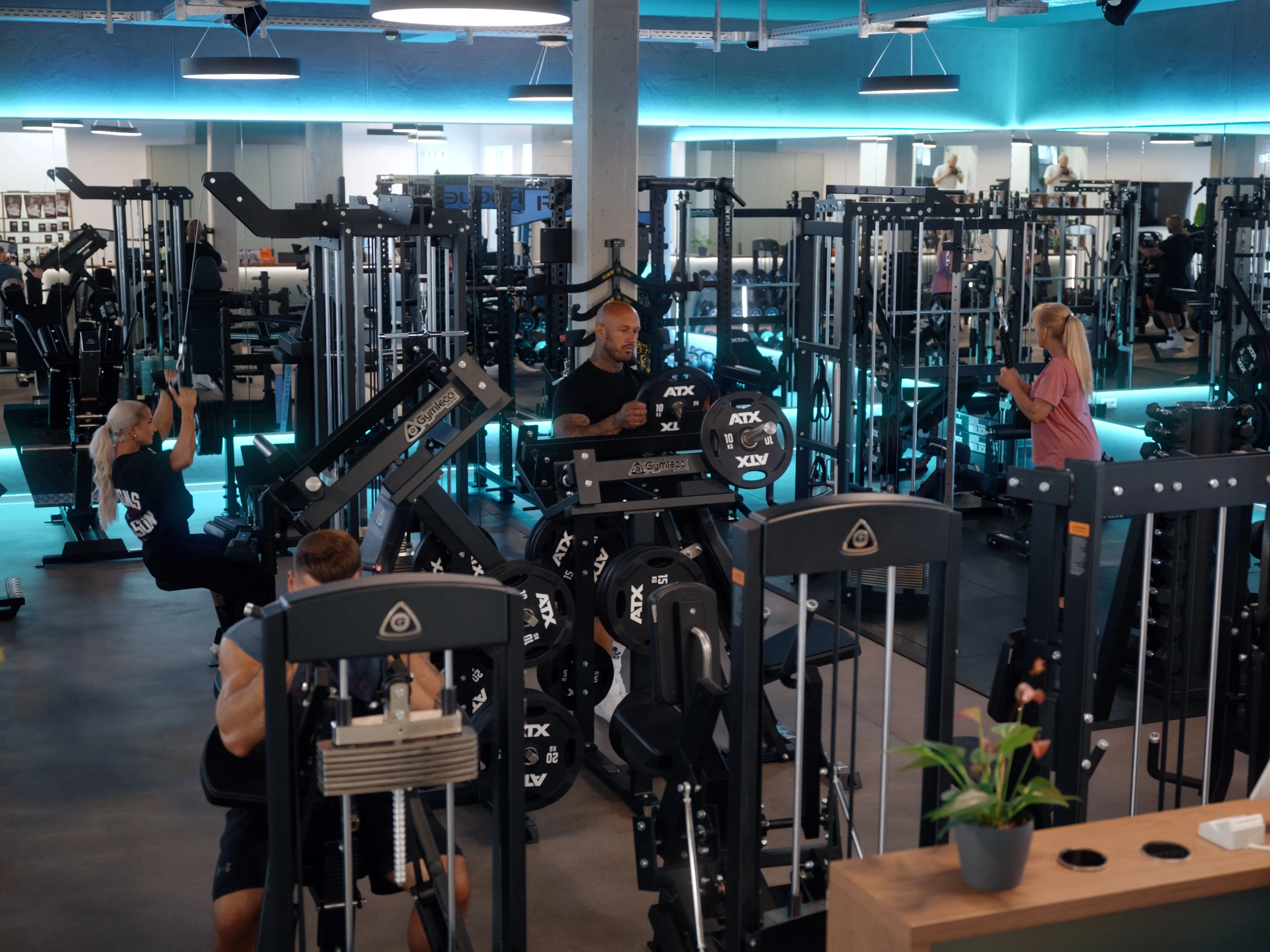 Menschen trainieren an verschiedenen Kraftgeräten in einem modernen Fitnessstudio mit Spiegeln und LED-Beleuchtung.