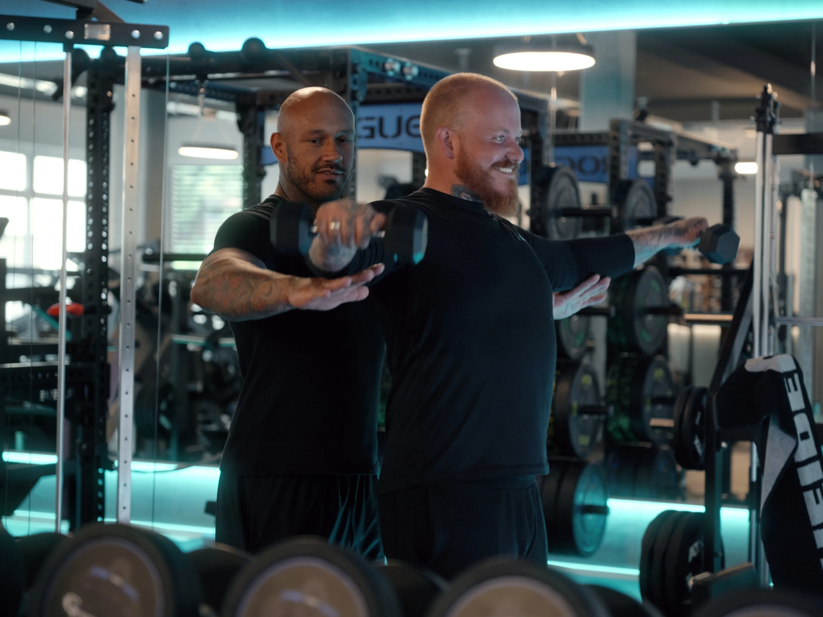 Trainer unterstützt Mann mit roten Bart, der im Fitnessstudio mit Kurzhanteln seitliche Armheben macht.