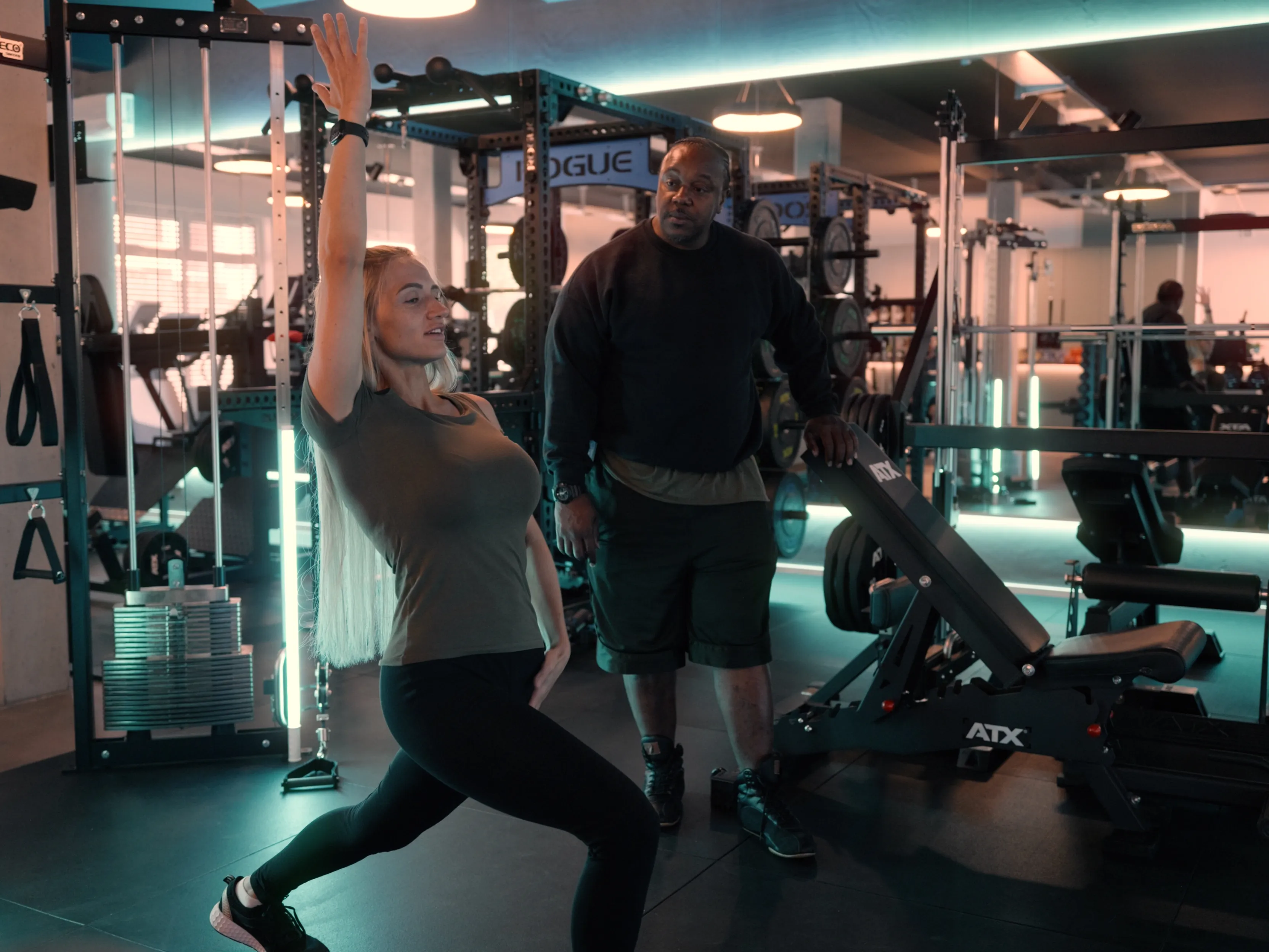 Frau macht Ausfallschritt im Fitnessstudio, während Trainer ihr zuschaut.