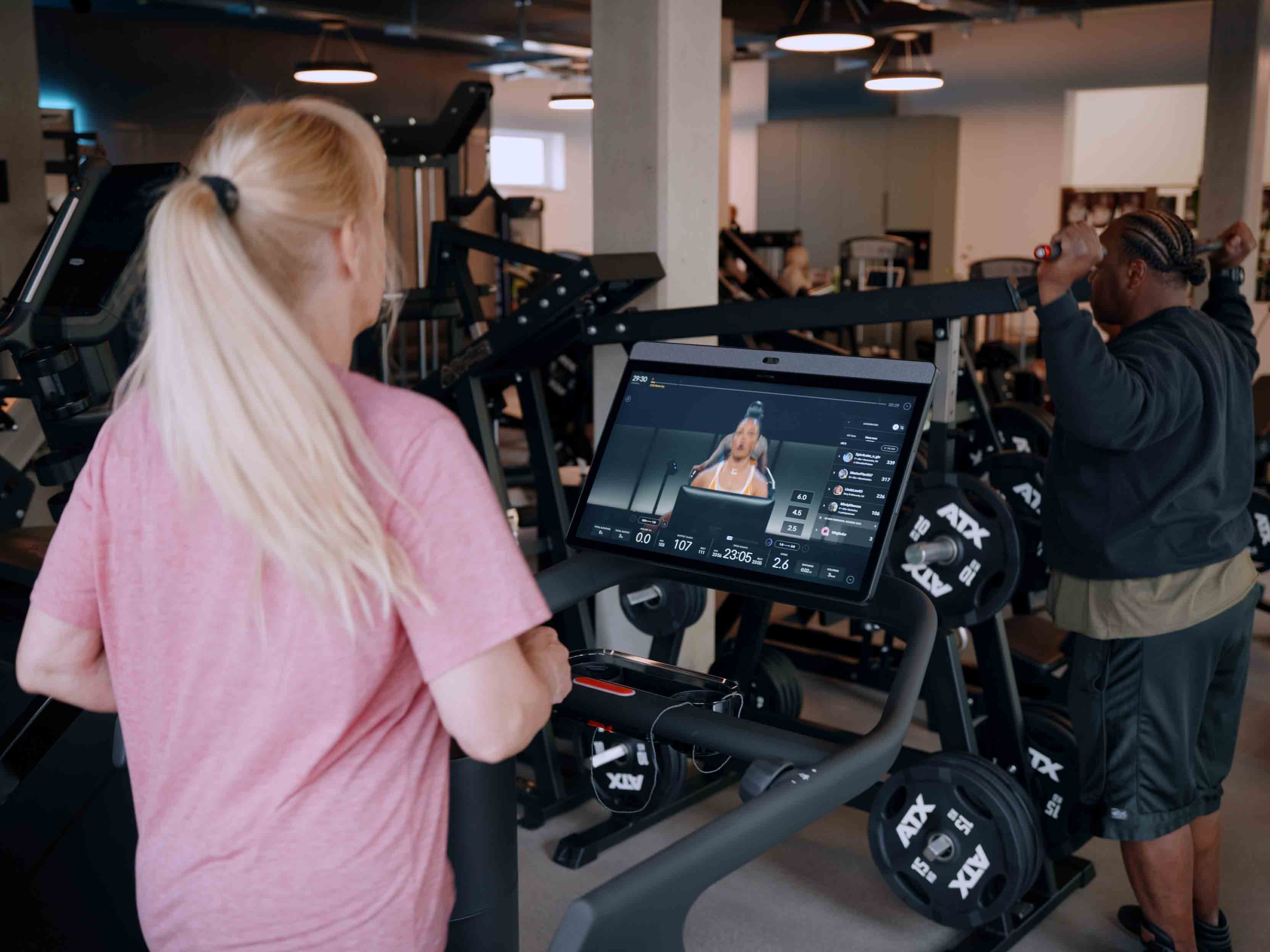 Frau auf Laufband mit Fitnessvideo auf Bildschirm, Mann trainiert mit Gewichten im Hintergrund.