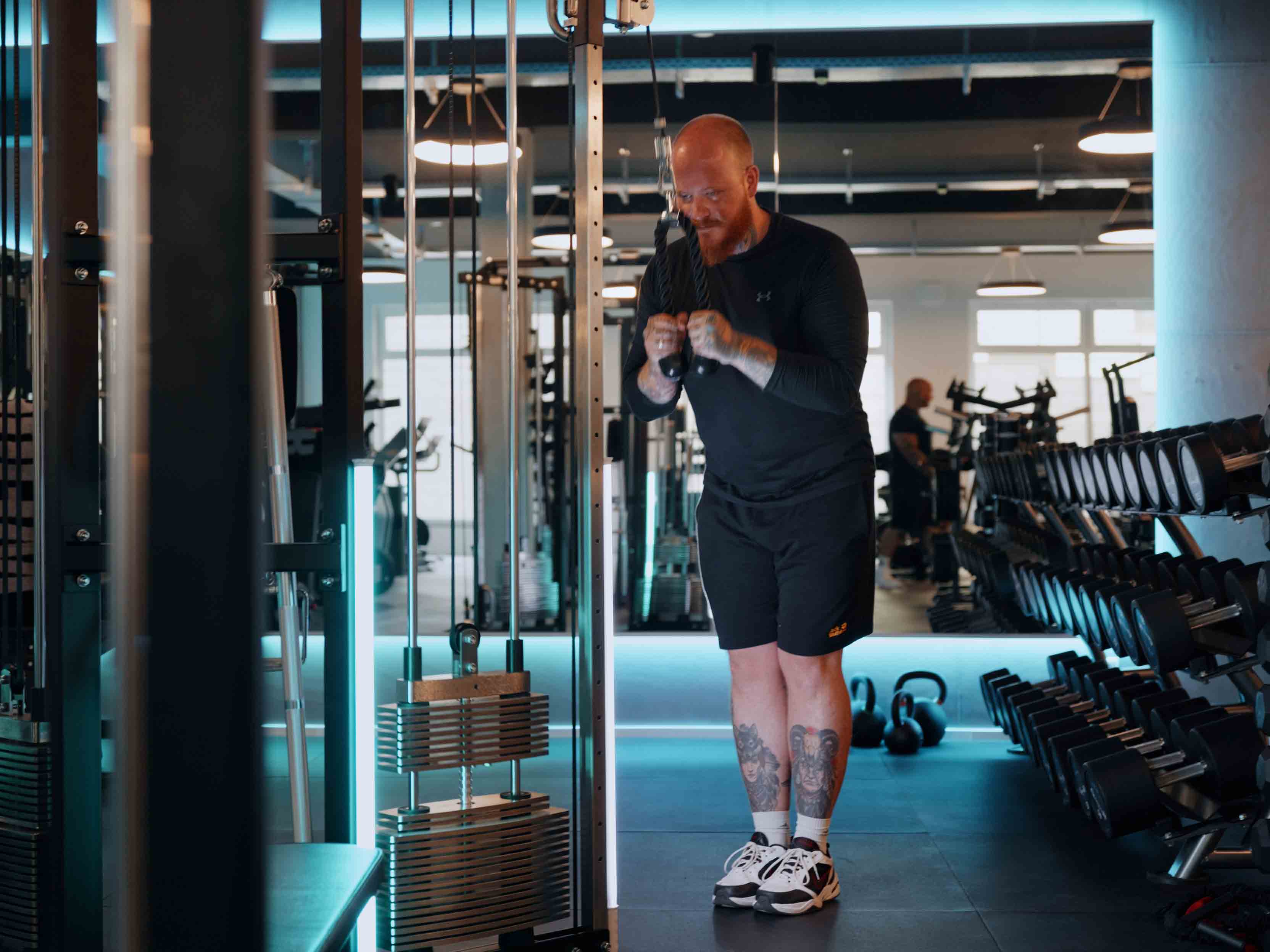 Mann trainiert Trizeps mit Kabelzugmaschine im Fitnessstudio, sichtbare Tattoos an den Beinen.