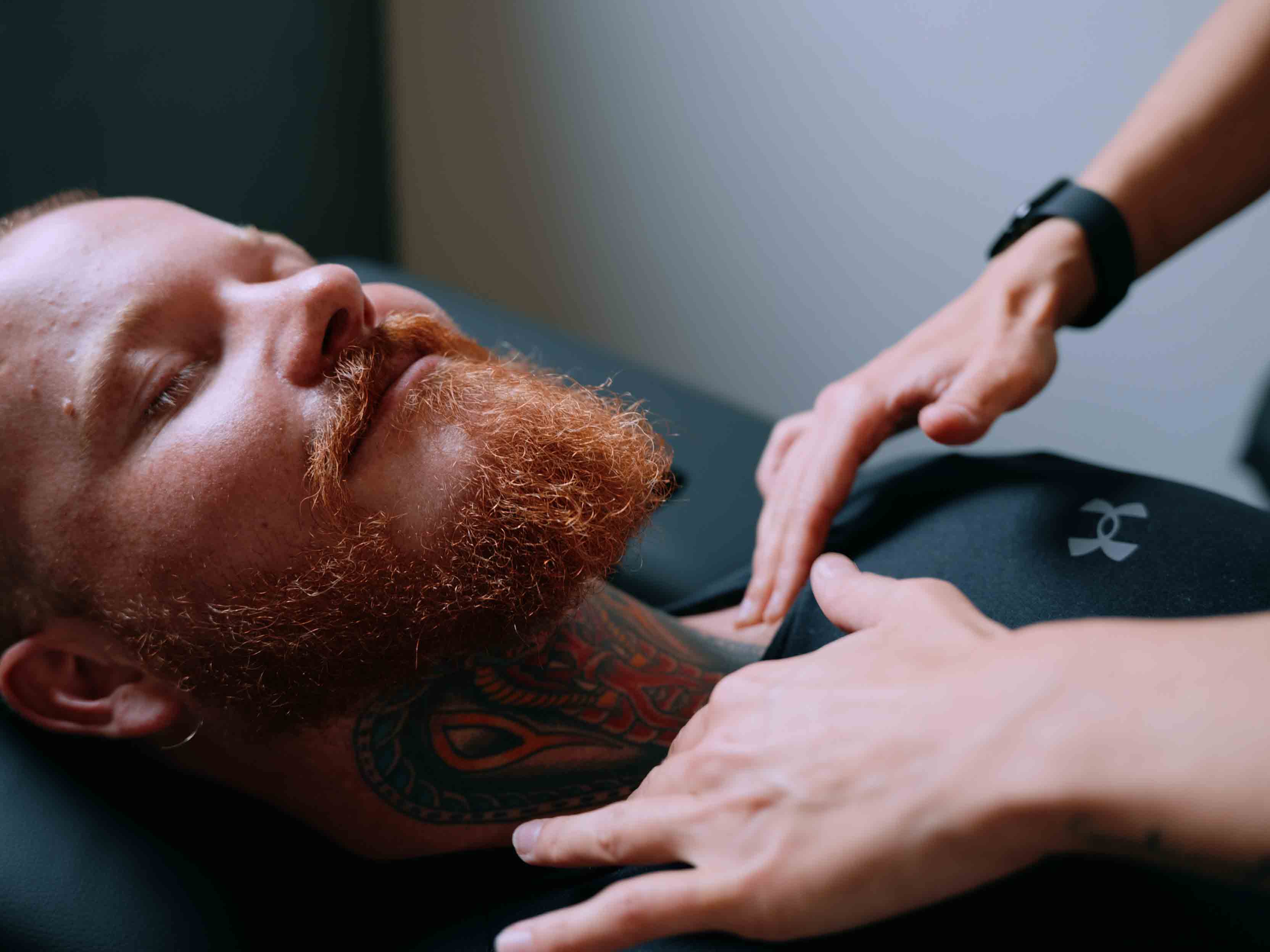 Mann mit rotem Bart und Hals-Tattoo liegt entspannt, Hände einer anderen Person sanft auf seiner Brust.