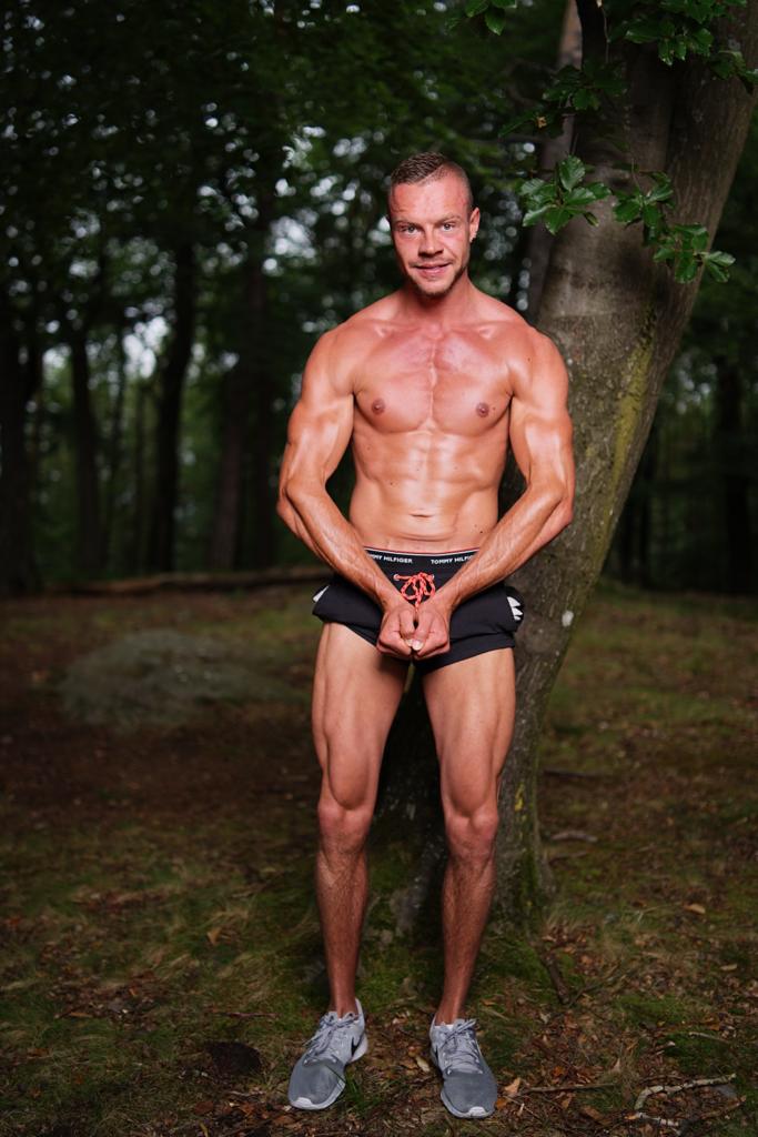 Muskelbepackter Mann in Shorts und Sportschuhen posiert vor einem Baum im Wald.