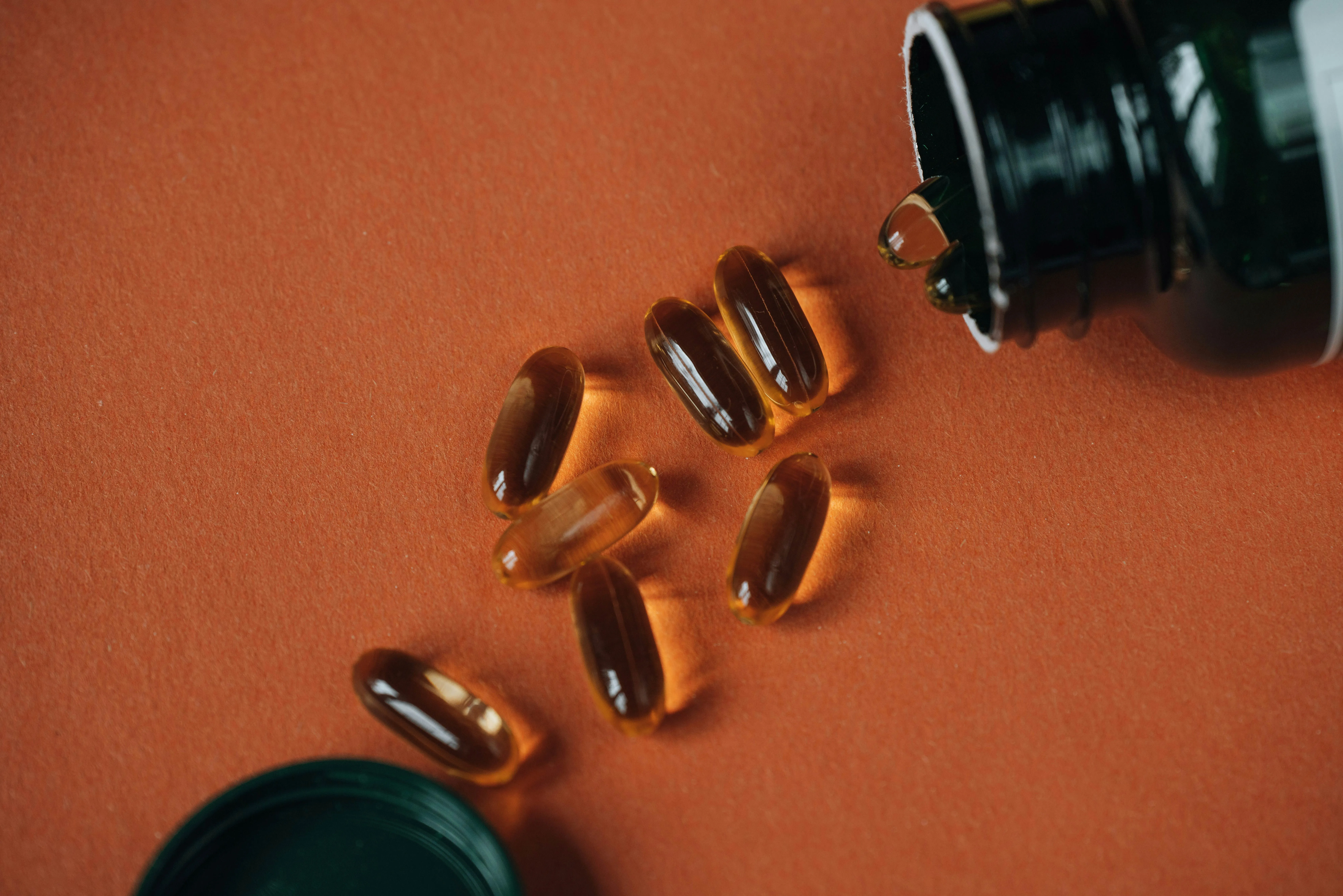 Transparente weiche Kapseln aus einem dunklen Fläschchen auf orangem Hintergrund.